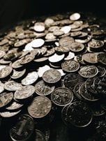 A large pile of coins is spread across a surface. The coins have varying designs and inscriptions, indicating they might be from different mints or countries. The lighting is dark with only a few reflections creating a metallic shine on some coins.