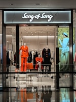 A modern retail store display featuring mannequins dressed in vibrant and fashionable clothing. The storefront includes elegant outfits in shades of orange and black with a stylish backdrop, and an illuminated sign reads 'Song of Song'. The interior appears sleek with a glossy floor and mirrored reflections.