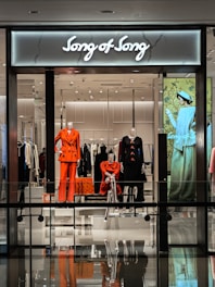 A modern retail store display featuring mannequins dressed in vibrant and fashionable clothing. The storefront includes elegant outfits in shades of orange and black with a stylish backdrop, and an illuminated sign reads 'Song of Song'. The interior appears sleek with a glossy floor and mirrored reflections.