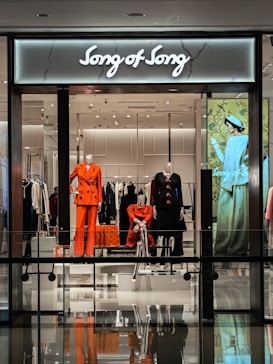 A modern retail store display featuring mannequins dressed in vibrant and fashionable clothing. The storefront includes elegant outfits in shades of orange and black with a stylish backdrop, and an illuminated sign reads 'Song of Song'. The interior appears sleek with a glossy floor and mirrored reflections.