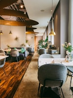 A modern, stylish restaurant interior with soft lighting and a warm ambiance. The space features several round tables with gray cushioned chairs, elegant plant decorations, and hanging pendant lights. The flooring combines polished wood and concrete textures, adding to the sophisticated design. Large windows on the right allow natural light to enter, complementing the inviting atmosphere. Thoughtfully placed vases with flowers enhance the aesthetic appeal.