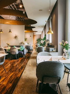A modern, stylish restaurant interior with soft lighting and a warm ambiance. The space features several round tables with gray cushioned chairs, elegant plant decorations, and hanging pendant lights. The flooring combines polished wood and concrete textures, adding to the sophisticated design. Large windows on the right allow natural light to enter, complementing the inviting atmosphere. Thoughtfully placed vases with flowers enhance the aesthetic appeal.