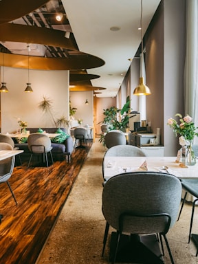 A modern, stylish restaurant interior with soft lighting and a warm ambiance. The space features several round tables with gray cushioned chairs, elegant plant decorations, and hanging pendant lights. The flooring combines polished wood and concrete textures, adding to the sophisticated design. Large windows on the right allow natural light to enter, complementing the inviting atmosphere. Thoughtfully placed vases with flowers enhance the aesthetic appeal.