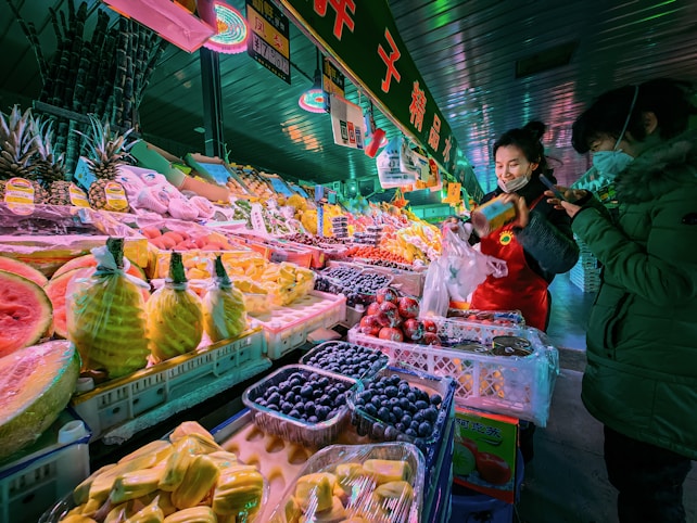 A vibrant market scene filled with an array of colorful fruits neatly arranged on stalls. Two people are interacting, one behind the counter wearing a red apron, possibly a vendor, and the other, possibly a customer, wearing a green jacket. The display includes pineapples, watermelon, blueberries, grapes, and other tropical fruits, with bright lighting enhancing their colors.