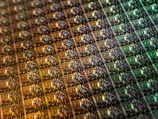 An array of intricate, patterned semiconductor chips with a reflective sheen, showing various colors and geometric patterns. The surface appears highly detailed with a grid-like structure.
