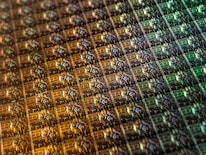 An array of intricate, patterned semiconductor chips with a reflective sheen, showing various colors and geometric patterns. The surface appears highly detailed with a grid-like structure.