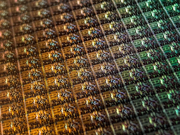 An array of intricate, patterned semiconductor chips with a reflective sheen, showing various colors and geometric patterns. The surface appears highly detailed with a grid-like structure.