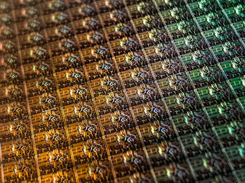 An array of intricate, patterned semiconductor chips with a reflective sheen, showing various colors and geometric patterns. The surface appears highly detailed with a grid-like structure.