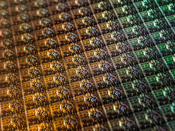 An array of intricate, patterned semiconductor chips with a reflective sheen, showing various colors and geometric patterns. The surface appears highly detailed with a grid-like structure.