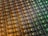 An array of intricate, patterned semiconductor chips with a reflective sheen, showing various colors and geometric patterns. The surface appears highly detailed with a grid-like structure.