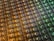 An array of intricate, patterned semiconductor chips with a reflective sheen, showing various colors and geometric patterns. The surface appears highly detailed with a grid-like structure.