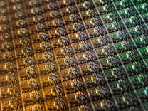 An array of intricate, patterned semiconductor chips with a reflective sheen, showing various colors and geometric patterns. The surface appears highly detailed with a grid-like structure.