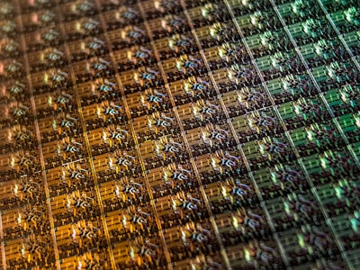An array of intricate, patterned semiconductor chips with a reflective sheen, showing various colors and geometric patterns. The surface appears highly detailed with a grid-like structure.