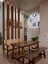 A cozy interior space featuring a rustic wooden table with matching benches. In the background, vertical brown columns and a partially exposed white brick wall add character to the room. On the wall, a bright sign reads 'GOOD VIBES ONLY,' enhancing the inviting atmosphere. There's a green potted plant and vintage posters creating a warm and casual ambiance.