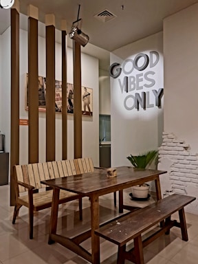 A cozy interior space featuring a rustic wooden table with matching benches. In the background, vertical brown columns and a partially exposed white brick wall add character to the room. On the wall, a bright sign reads 'GOOD VIBES ONLY,' enhancing the inviting atmosphere. There's a green potted plant and vintage posters creating a warm and casual ambiance.