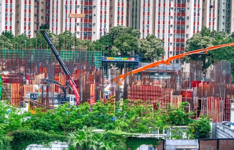 A bustling construction site surrounded by greenery, with metal beams and columns in various stages of assembly. Two large cranes, one black and another orange, are positioned prominently, aiding in the construction efforts. In the background, a residential building with red and white patterns is visible beyond a line of trees.