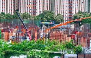 A bustling construction site surrounded by greenery, with metal beams and columns in various stages of assembly. Two large cranes, one black and another orange, are positioned prominently, aiding in the construction efforts. In the background, a residential building with red and white patterns is visible beyond a line of trees.