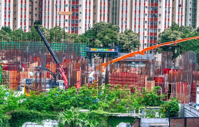 A bustling construction site surrounded by greenery, with metal beams and columns in various stages of assembly. Two large cranes, one black and another orange, are positioned prominently, aiding in the construction efforts. In the background, a residential building with red and white patterns is visible beyond a line of trees.