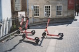 Three electric scooters are parked on a paved street near a building with old-style architecture. The building has large windows and a red sign indicating a museum. There is a black iron fence on the left and a path leading away from the area.