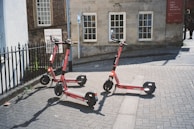 Three electric scooters are parked on a paved street near a building with old-style architecture. The building has large windows and a red sign indicating a museum. There is a black iron fence on the left and a path leading away from the area.
