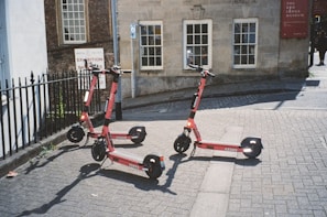Three electric scooters are parked on a paved street near a building with old-style architecture. The building has large windows and a red sign indicating a museum. There is a black iron fence on the left and a path leading away from the area.