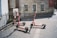 Three electric scooters are parked on a paved street near a building with old-style architecture. The building has large windows and a red sign indicating a museum. There is a black iron fence on the left and a path leading away from the area.