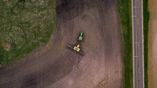 An aerial view of a tractor on agricultural land, cultivating a field with visible tire tracks and variations in soil color. Adjacent to the field is a paved road bordered by grass, with a distinct contrast between the cultivated and uncultivated areas.