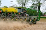 A large green and yellow agricultural machine is being operated in a field. The machinery appears to be a planter or seeder, preparing the soil for planting crops. Dust is being kicked up from the ground as it moves through the field, indicating active farming activity. In the background, there are trees and a red barn can be seen.