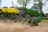 A large green and yellow agricultural machine is being operated in a field. The machinery appears to be a planter or seeder, preparing the soil for planting crops. Dust is being kicked up from the ground as it moves through the field, indicating active farming activity. In the background, there are trees and a red barn can be seen.