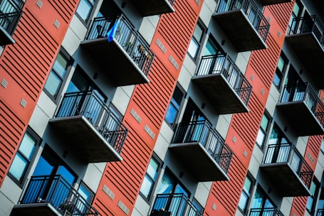 A modern apartment building facade with multiple levels of balconies jutting out. The structure features alternating orange panels and grey concrete sections. Each balcony is equipped with a black railing, and various plants and household items are visible. The windows are reflective, capturing the sky and surroundings.