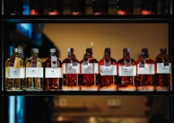 A row of glass bottles containing liquid, each with a label featuring text and icons. The liquids appear in shades of amber and yellow, and each bottle has a metal cap and a hanging tag with additional information. The bottles are aligned neatly on a dark, industrial-style shelf with a warm light in the background.