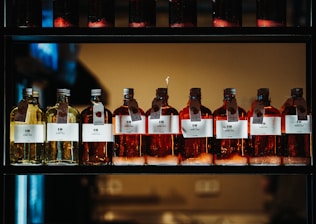 A row of glass bottles containing liquid, each with a label featuring text and icons. The liquids appear in shades of amber and yellow, and each bottle has a metal cap and a hanging tag with additional information. The bottles are aligned neatly on a dark, industrial-style shelf with a warm light in the background.