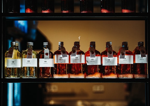 A row of glass bottles containing liquid, each with a label featuring text and icons. The liquids appear in shades of amber and yellow, and each bottle has a metal cap and a hanging tag with additional information. The bottles are aligned neatly on a dark, industrial-style shelf with a warm light in the background.