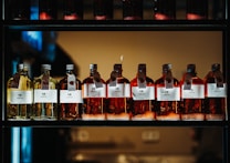 A row of glass bottles containing liquid, each with a label featuring text and icons. The liquids appear in shades of amber and yellow, and each bottle has a metal cap and a hanging tag with additional information. The bottles are aligned neatly on a dark, industrial-style shelf with a warm light in the background.