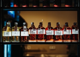 A row of glass bottles containing liquid, each with a label featuring text and icons. The liquids appear in shades of amber and yellow, and each bottle has a metal cap and a hanging tag with additional information. The bottles are aligned neatly on a dark, industrial-style shelf with a warm light in the background.