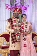A couple is dressed in traditional Indian wedding attire, standing in front of a decorated backdrop with the names 'Raksh' and 'Ajay'. The man wears a red and gold sherwani with a turban and the woman wears a pink embellished lehenga. Both are adorned with floral garlands.