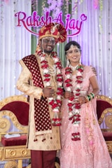 A couple is dressed in traditional Indian wedding attire, standing in front of a decorated backdrop with the names 'Raksh' and 'Ajay'. The man wears a red and gold sherwani with a turban and the woman wears a pink embellished lehenga. Both are adorned with floral garlands.