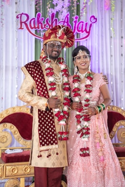 A couple is dressed in traditional Indian wedding attire, standing in front of a decorated backdrop with the names 'Raksh' and 'Ajay'. The man wears a red and gold sherwani with a turban and the woman wears a pink embellished lehenga. Both are adorned with floral garlands.