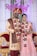 A couple is dressed in traditional Indian wedding attire, standing in front of a decorated backdrop with the names 'Raksh' and 'Ajay'. The man wears a red and gold sherwani with a turban and the woman wears a pink embellished lehenga. Both are adorned with floral garlands.