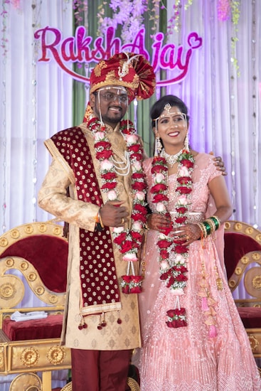 A couple is dressed in traditional Indian wedding attire, standing in front of a decorated backdrop with the names 'Raksh' and 'Ajay'. The man wears a red and gold sherwani with a turban and the woman wears a pink embellished lehenga. Both are adorned with floral garlands.