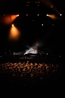 A concert scene with a large audience silhouetted against stage lights. The stage is illuminated with dramatic lighting, focusing on a drum set. Warm lights create a vibrant atmosphere, highlighting the crowd's excitement.