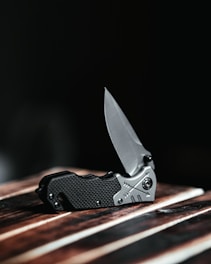 A sleek folding knife with a metallic blade and textured handle rests on a wooden surface. The blade is partially open and glistens under subdued lighting, casting a sharp and polished appearance. The dark background accentuates the knife's details and craftsmanship.