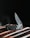 A sleek folding knife with a metallic blade and textured handle rests on a wooden surface. The blade is partially open and glistens under subdued lighting, casting a sharp and polished appearance. The dark background accentuates the knife's details and craftsmanship.