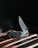 A sleek folding knife with a metallic blade and textured handle rests on a wooden surface. The blade is partially open and glistens under subdued lighting, casting a sharp and polished appearance. The dark background accentuates the knife's details and craftsmanship.