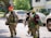 Three firefighters wearing full gear and yellow helmets walk down a street past parked vehicles. One firefighter carries a hose while another carries an axe. Road signs are visible, and lush greenery surrounds the area.