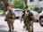 Three firefighters wearing full gear and yellow helmets walk down a street past parked vehicles. One firefighter carries a hose while another carries an axe. Road signs are visible, and lush greenery surrounds the area.
