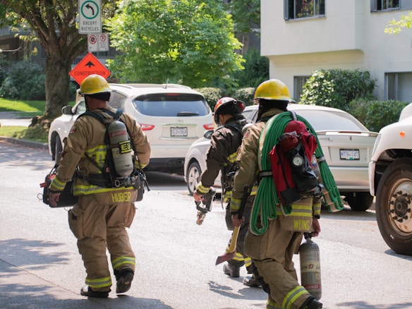 Three firefighters wearing full gear and yellow helmets walk down a street past parked vehicles. One firefighter carries a hose while another carries an axe. Road signs are visible, and lush greenery surrounds the area.
