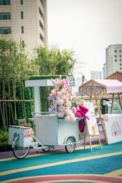 A decorative white ice cream cart adorned with a vibrant bouquet of flowers and a large pink ribbon. The cart is positioned on a colorful, circular-patterned floor with bamboo plants and a tall building in the background. Nearby, there is a whiteboard with information and a tent-style cover.
