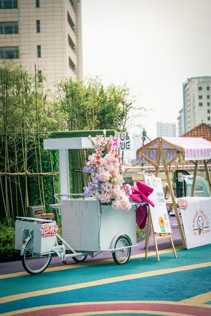 A decorative white ice cream cart adorned with a vibrant bouquet of flowers and a large pink ribbon. The cart is positioned on a colorful, circular-patterned floor with bamboo plants and a tall building in the background. Nearby, there is a whiteboard with information and a tent-style cover.