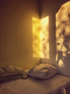 Soft, warm light creates a shadow pattern on the wall, revealing the silhouette of a person holding a camera. A simple bed with a pillow and rumpled blanket is positioned against the corner, suggesting a cozy and serene atmosphere.
